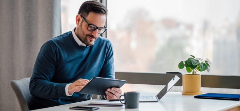 Man recherchiert mit Brille und blauem Pulli am Tablet zur Generationsnachfolge für Ärzte, sitzt am Schreibtisch.