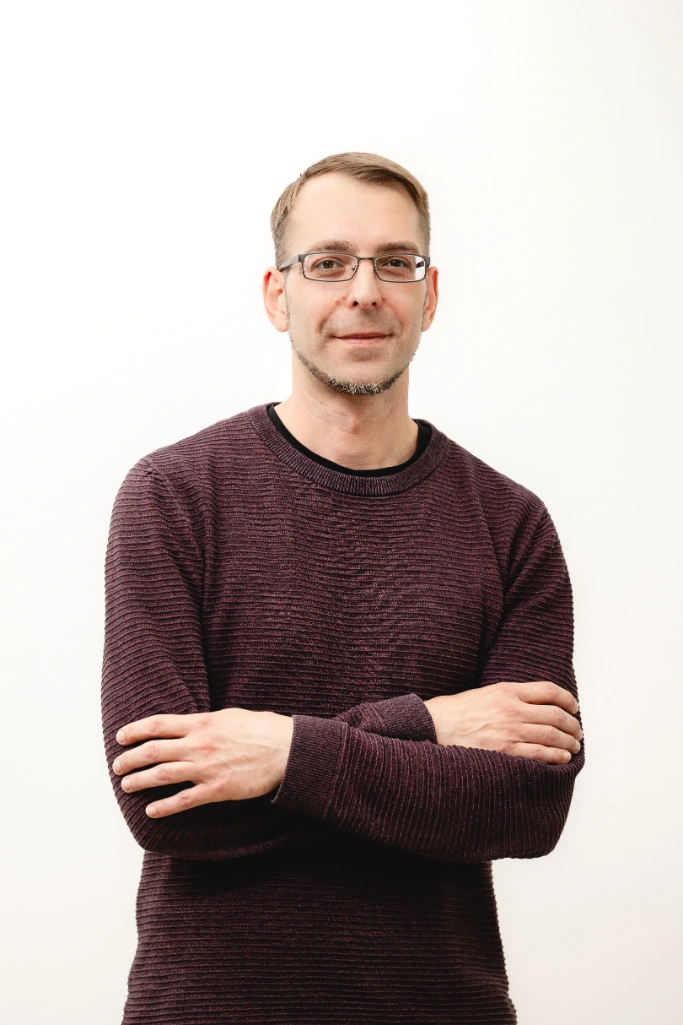 Mann mit kurzen braunen Haaren und Brille, im kastanienbraunen Pullover, verschränkt die Arme und lächelt leicht vor einem schlichten weißen Hintergrund.