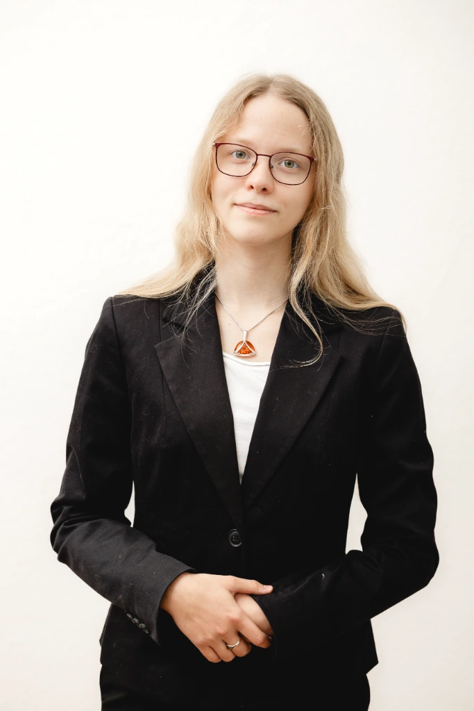 Junge blonde Frau mit Brille in schwarzem Blazer und weißem Hemd, steht vor weißem Hintergrund und schaut in die Kamera.