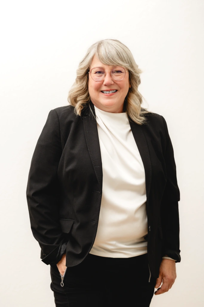 Eine lächelnde Frau mit grauem schulterlangem Haar, Brille, schwarzem Blazer und weißer Bluse steht vor einem schlichten weißen Hintergrund.