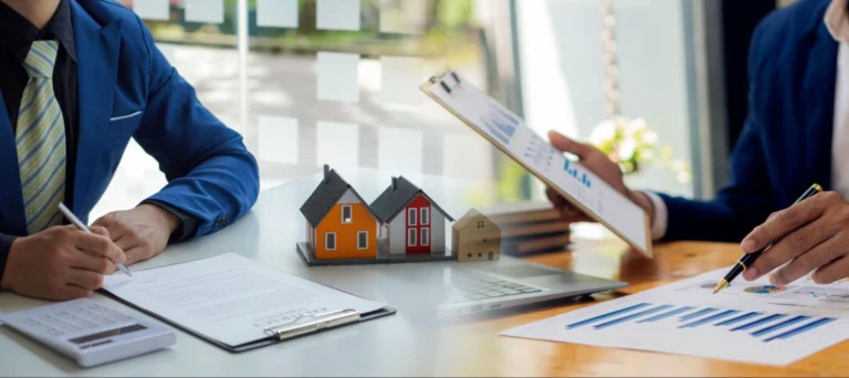 Zwei Geschäftsleute besprechen am Schreibtisch mit Hausmodellen, Diagrammen und Klemmbrett vermutlich Immobilienfragen.