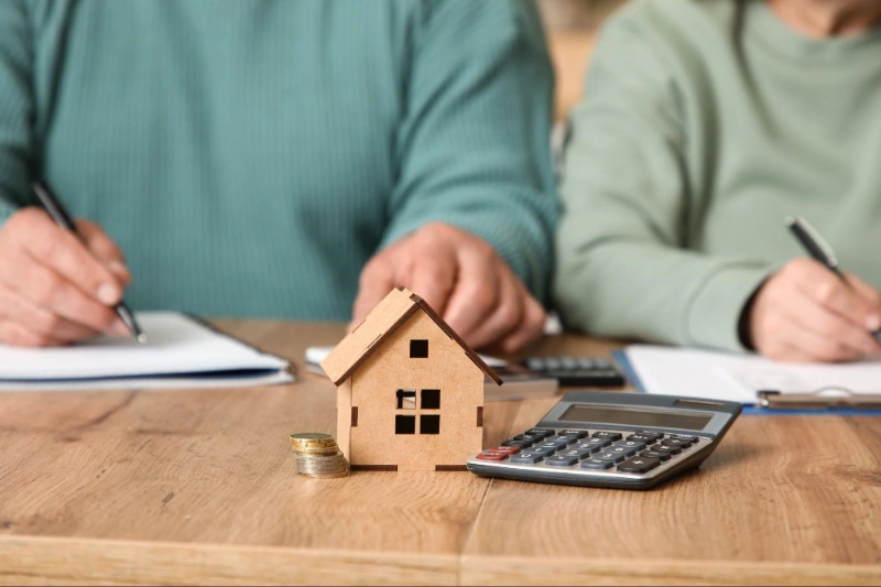 Holzhausmodell, Münzen und Taschenrechner auf einem Tisch; im Hintergrund schreiben zwei Personen in Grün.